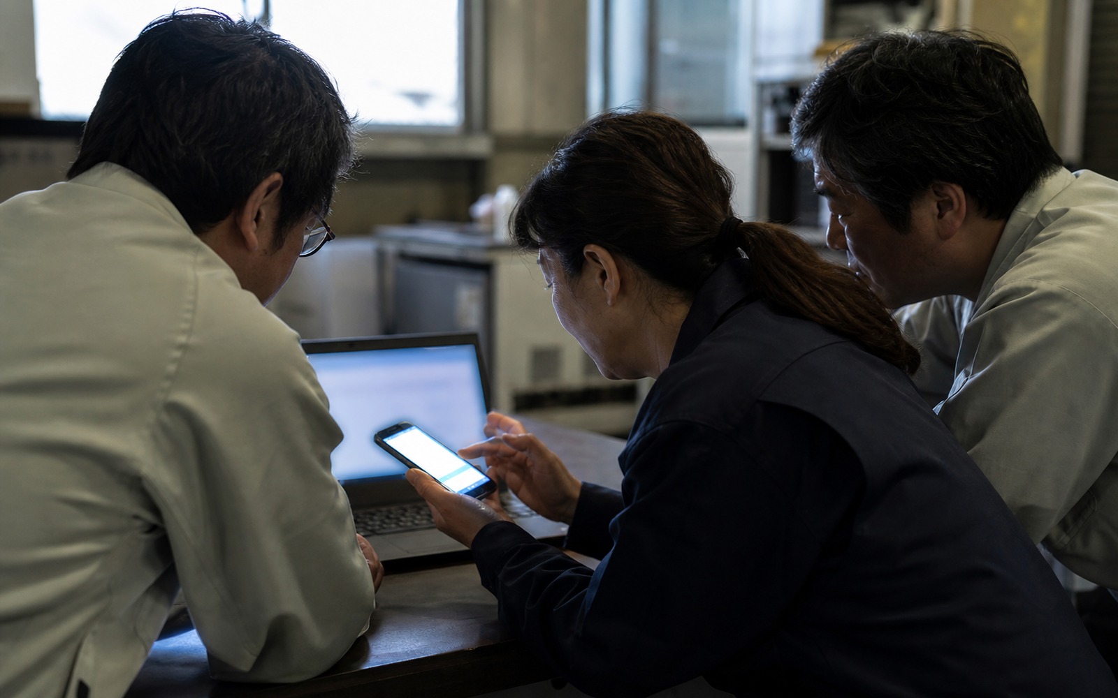 工場の作業場で、スマートフォンの画面を三人で覗き込んでいる様子